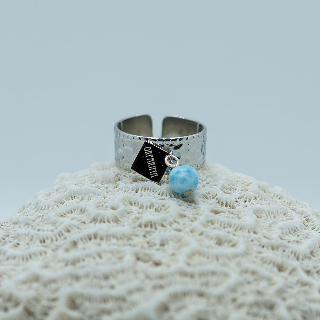 Bague texturé tendance de couleur argentée posée sur un coquillage. Bague en acier inoxydable 316L avec une breloque et une perle de larimar. Bague réglable adaptable à toutes les tailes de doigts.