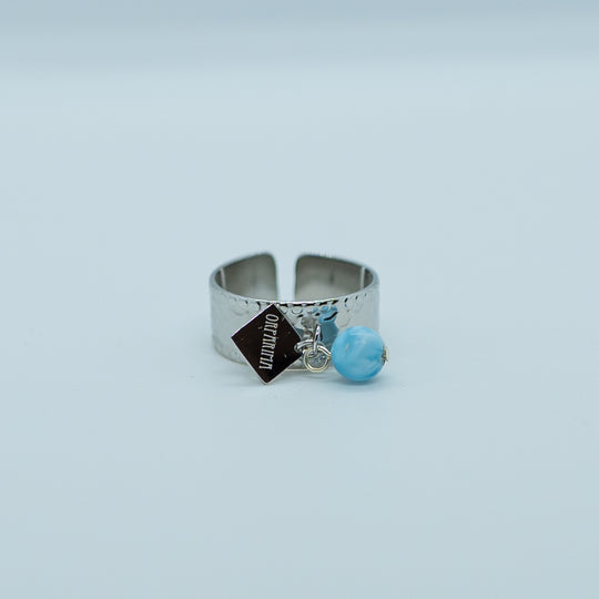 Bague ouverte réglable en acier inoxydable chirurgical 316L couleur argent. Bague texturée avec un anneau sur le heut de la bague ou sont accrochés une perle de larimar de couleur bleue et une breloque Orparima.