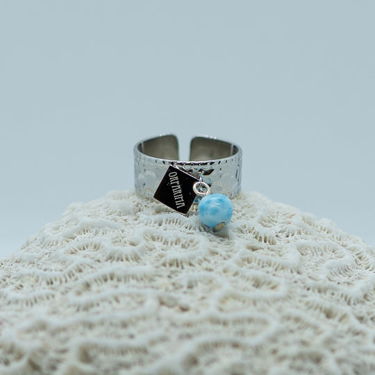 Bague texturé tendance de couleur argentée posée sur un coquillage. Bague en acier inoxydable 316L avec une breloque et une perle de larimar. Bague réglable adaptable à toutes les tailes de doigts.