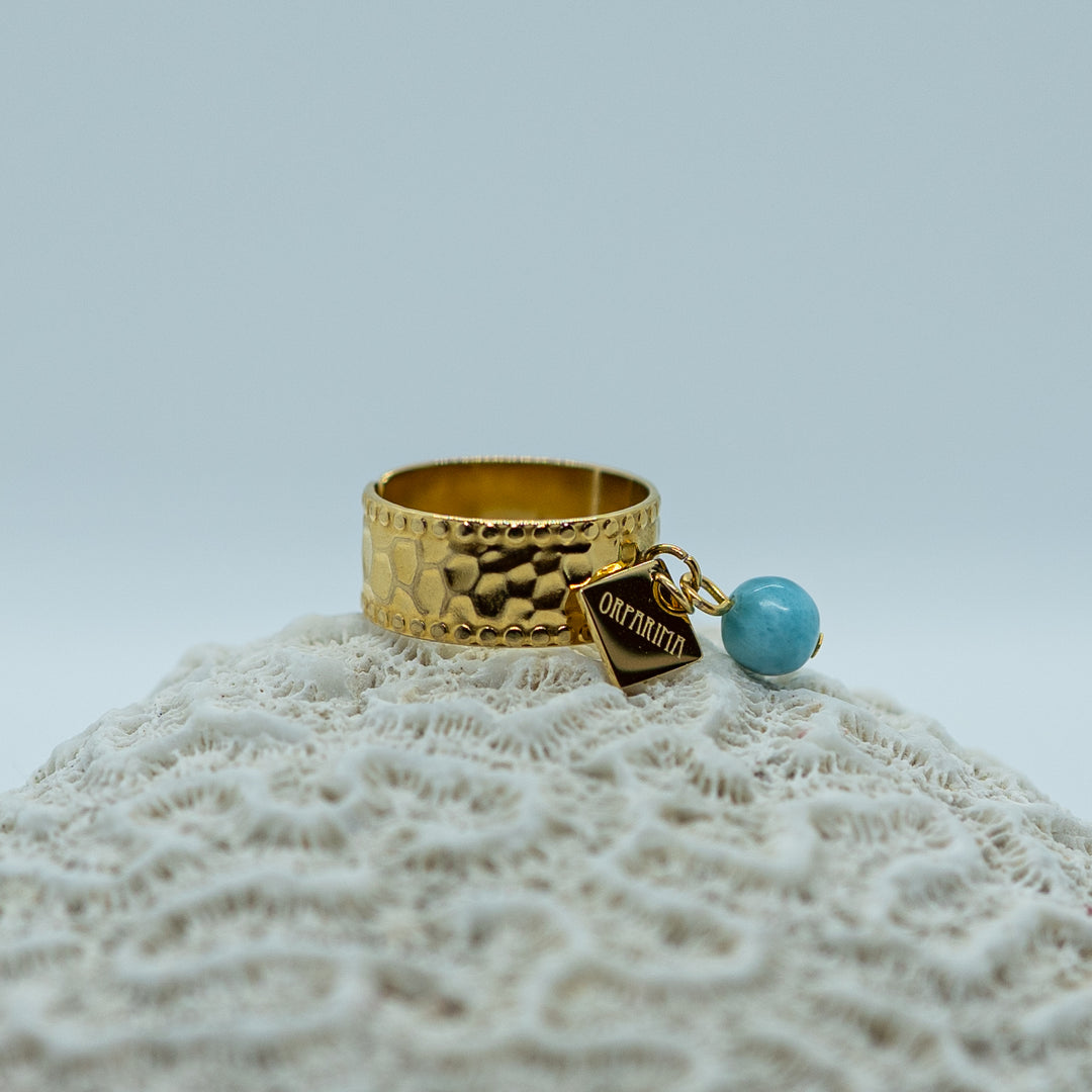 Bague en acier inoxydable 316L de couleur dorée posé sur un coquillage. Une breloque Orparima et une perle de larimar bleu sont accrochés à l&
