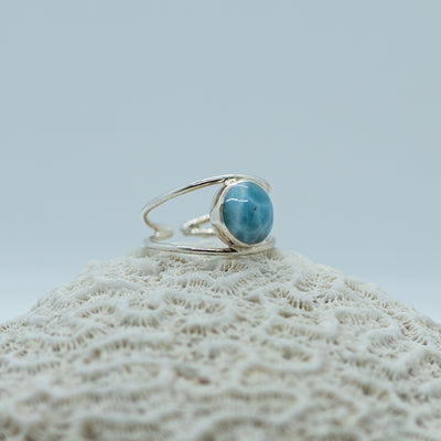 Bague posée de coté sur un coquillage sur fond blanc. Bague ornée d'une pierre de larimar de forme ronde. Bague non fermée réglable.