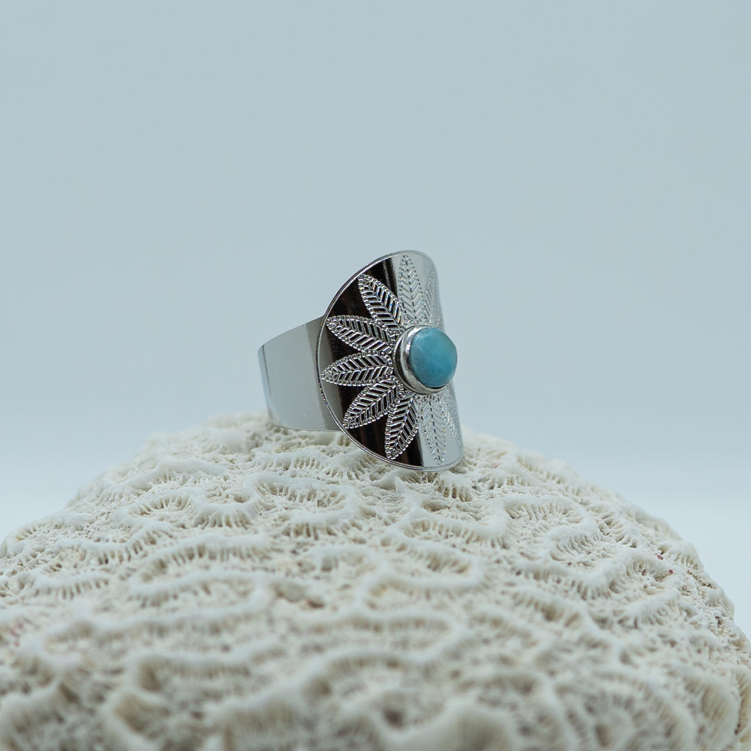 Bague originale en acier inoxydable 316L chirurgical de couleur argentée posée sur un coquillage sur fond blanc. Bague ouverte en dessous pour être réglable avec une pierre de larimar au centre de la bague.