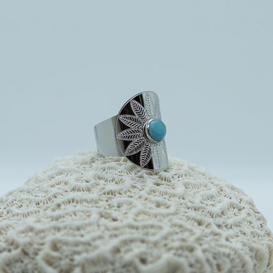 Bague originale en acier inoxydable 316L chirurgical de couleur argentée posée sur un coquillage sur fond blanc. Bague ouverte en dessous pour être réglable avec une pierre de larimar au centre de la bague.