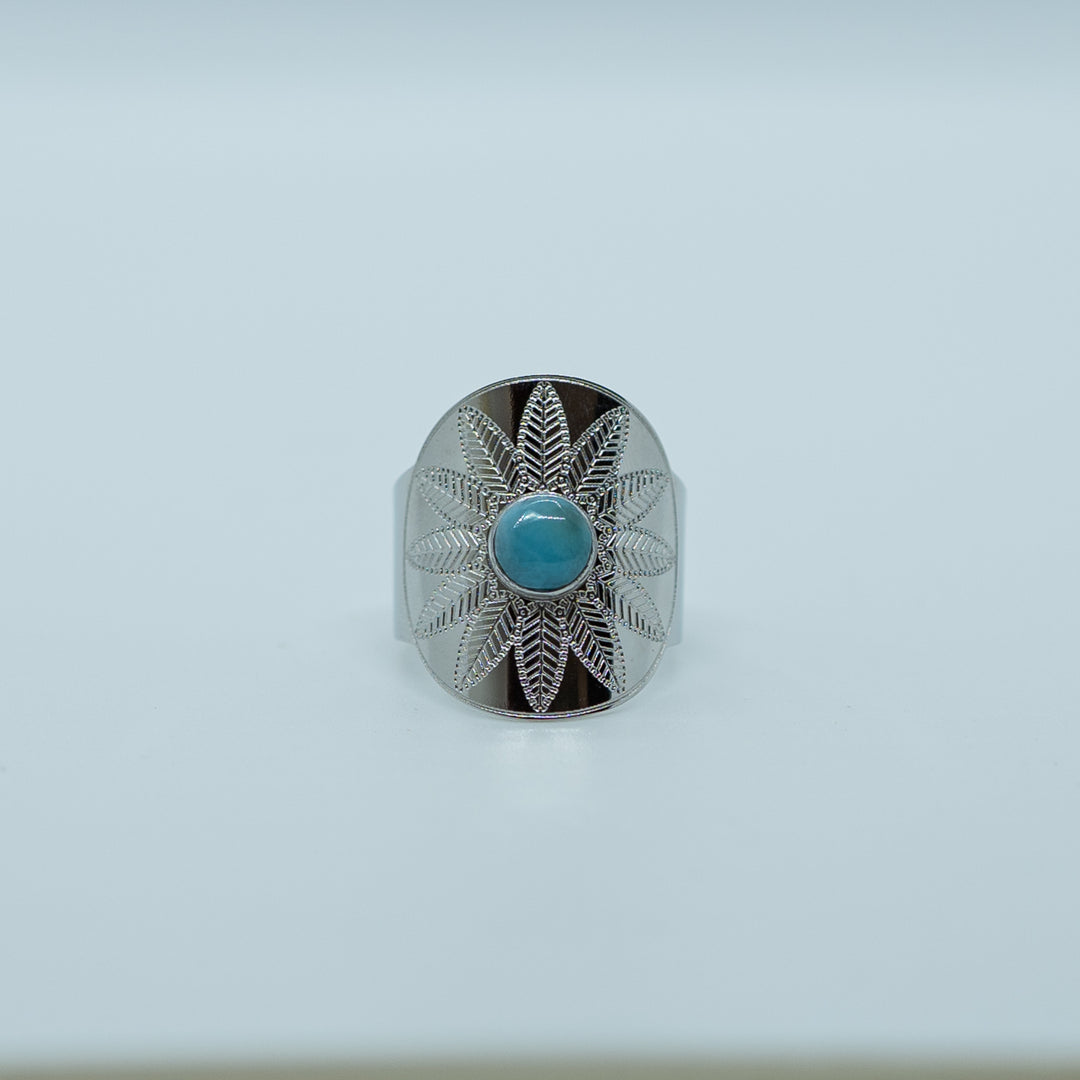 Bague neiba avec une gravure de fleur, sur fond blanc, avec une pierre de larimar au centre de la bague. Matériaux de fabrication acier inoxydable 316L argenté.Larimar de couleur bleu. Bague réglable.
