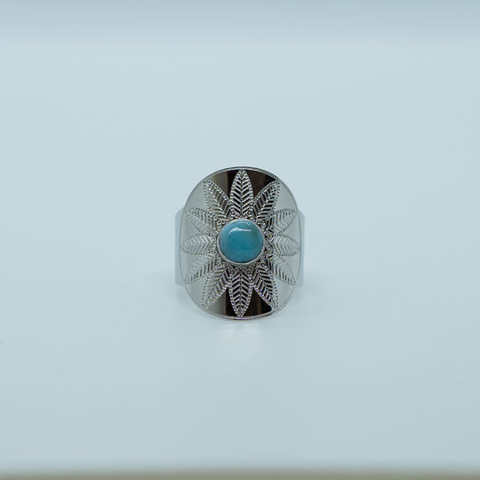 Bague neiba avec une gravure de fleur, sur fond blanc, avec une pierre de larimar au centre de la bague. Matériaux de fabrication acier inoxydable 316L argenté.Larimar de couleur bleu. Bague réglable.