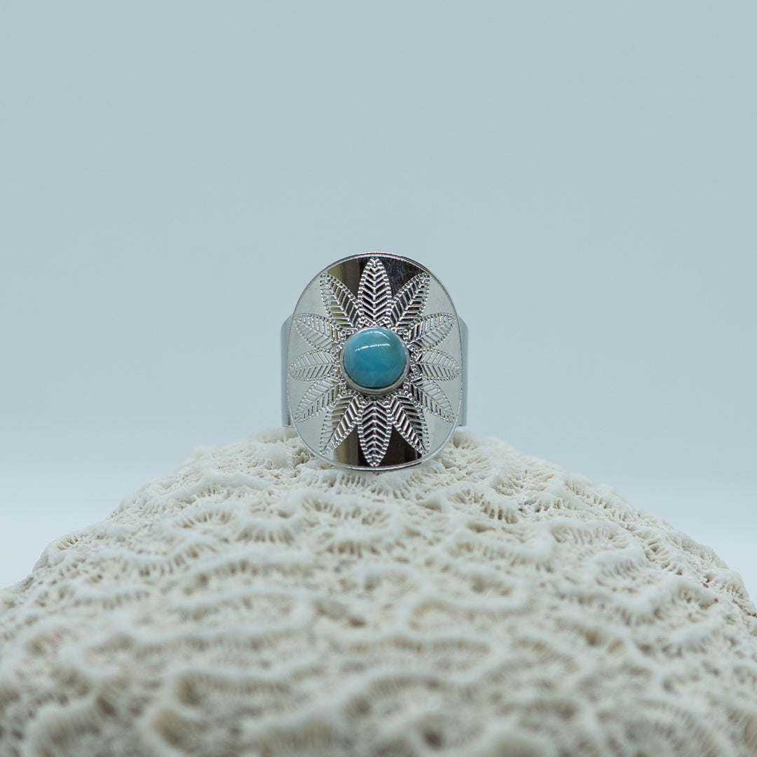 Superbe bague argentée en acier inoxydable 316L posée sur un coquillage, sur fond blanc. Perle de larimar bleu au centre de la bague. Gravure en forme de fleur.