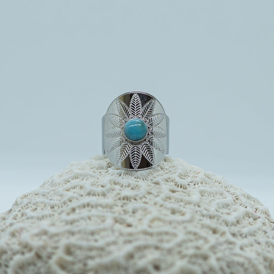 Superbe bague argentée en acier inoxydable 316L posée sur un coquillage, sur fond blanc. Perle de larimar bleu au centre de la bague. Gravure en forme de fleur.