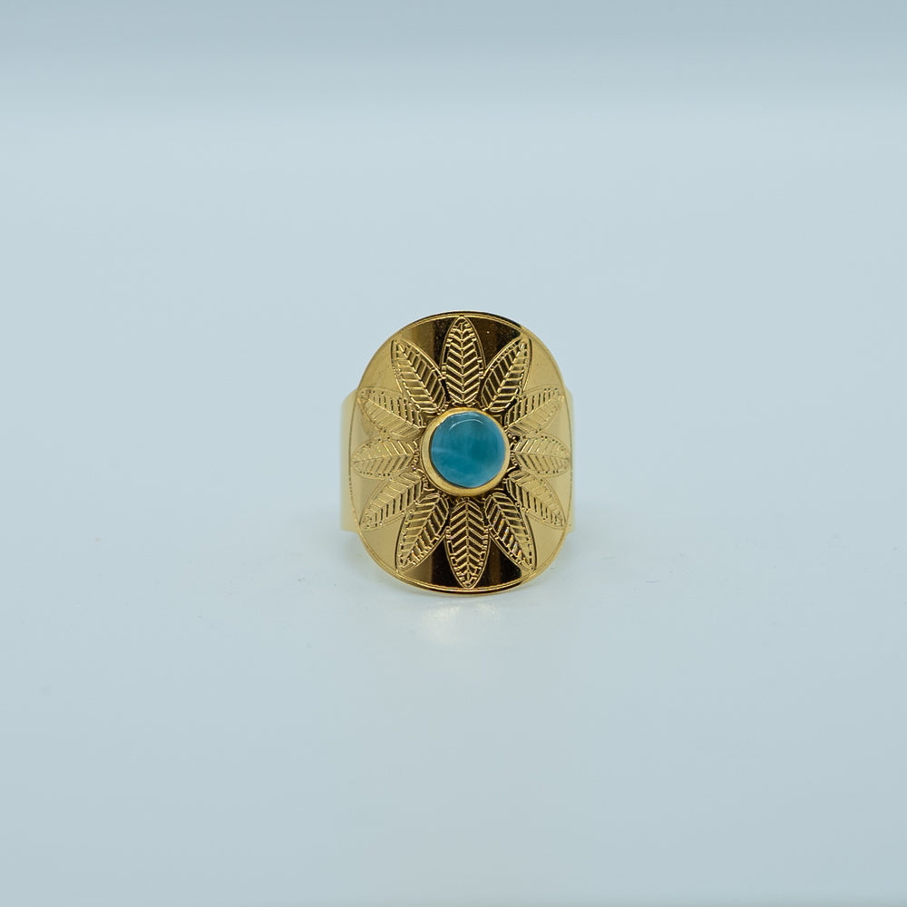 Bague dorée de face sur fond blanc, avec un cabochon de forme ronde collé en son centre. Bague avec une gravure en forme de fleur, ouverte derrière pour avoir différente tailles possible.