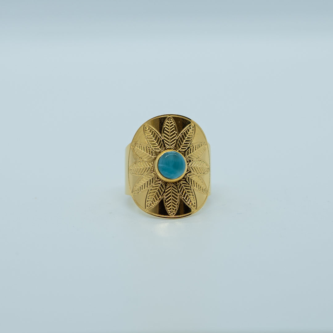 Bague dorée de face sur fond blanc, avec un cabochon de forme ronde collé en son centre. Bague avec une gravure en forme de fleur, ouverte derrière pour avoir différente tailles possible.