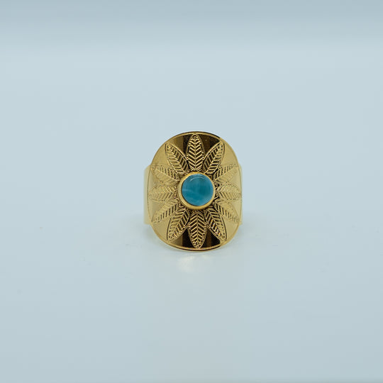 Bague dorée de face sur fond blanc, avec un cabochon de forme ronde collé en son centre. Bague avec une gravure en forme de fleur, ouverte derrière pour avoir différente tailles possible.