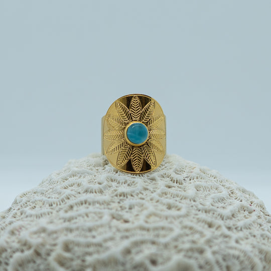 Bague de forme ovale avec une fleur gravé sur le dessus et une pierre de larimar ronde sertie en son centre. Bague en acier inoxydable 316L chirurgical qui ne ternie pas.