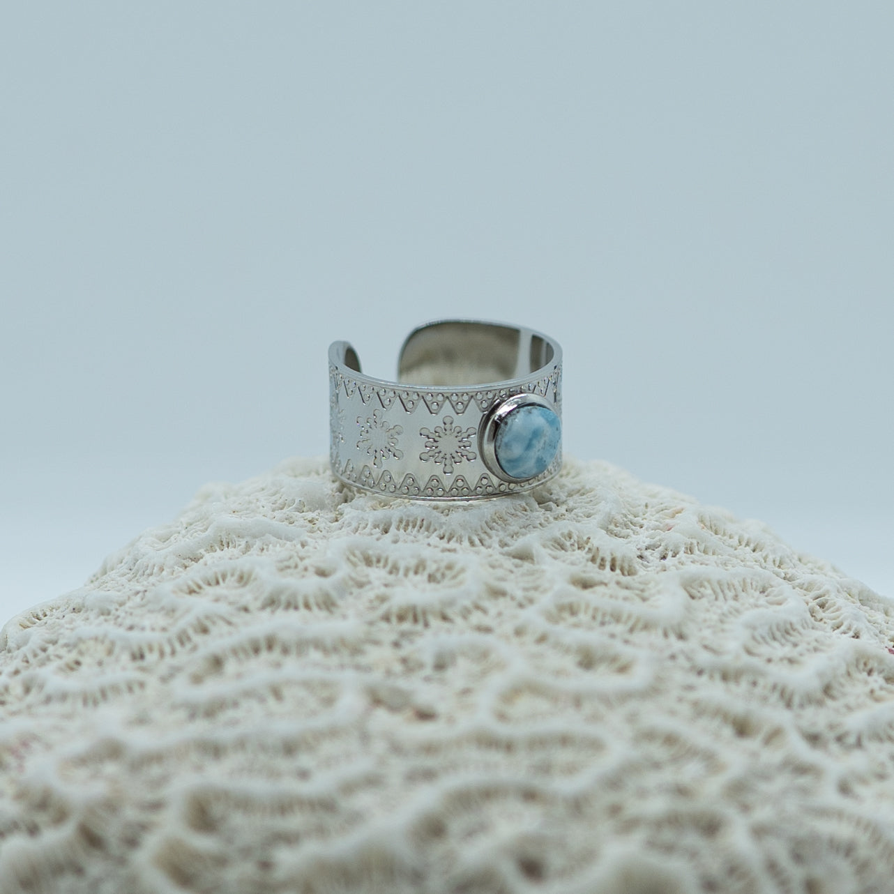 Bague posée de coté sur un coquillage. Bague en acier inoxydable chirurgical 316L couleur argent, ouverte pour se régler à plusieurs taille de doigts.