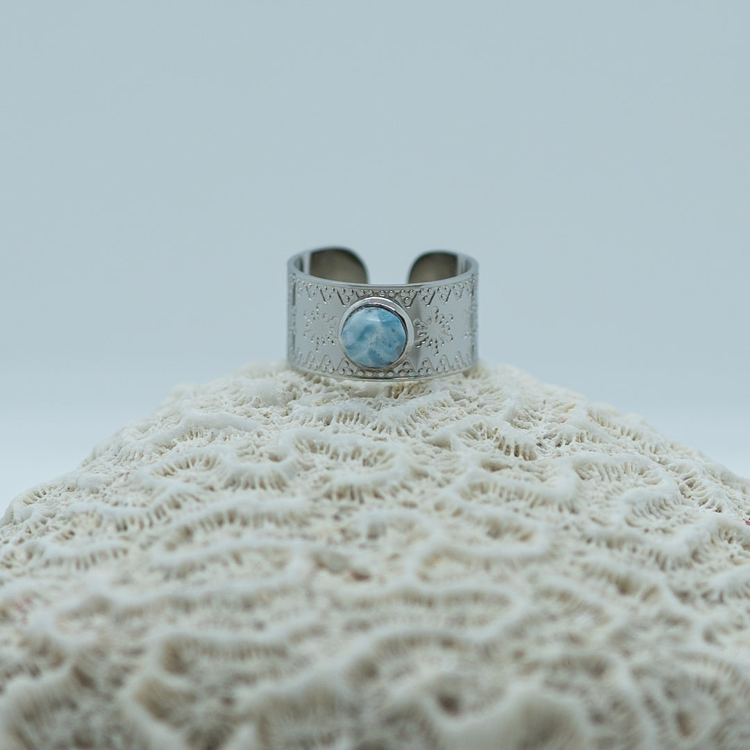 Bague avec une pierre de larimar en son centre. Bague posée sur un coquillage. Bijou avec des flocons de neige gravés sur le pourtour. Bague réglable couleur argent