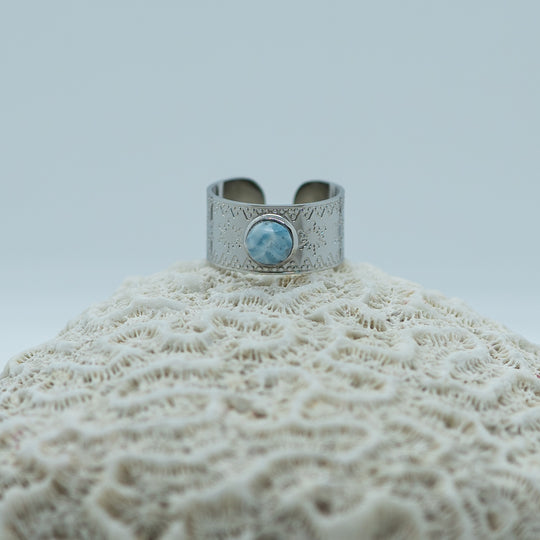 Bague avec une pierre de larimar en son centre. Bague posée sur un coquillage. Bijou avec des flocons de neige gravés sur le pourtour. Bague réglable couleur argent