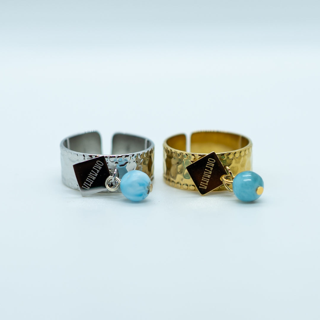 Deux bagues de face, posées sur le coté, texturées et réglables. Bagues faites en acier inoxydable chirurgical 316L, la bague de gauche est argenté et la bague de droite est dorée. Les deux bagues ont une breloque orparima et une perle de larimar attachées à un anneau.