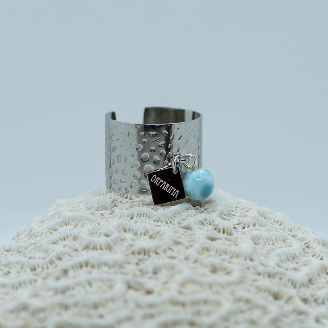 Bague couleur argent, en acier inoxydable 316L, posé sur un coquillage sur fond blanc. Bague avec une breloque orparima et une perle de 6mm en larimar pendant. Bague réglable et ajustable.