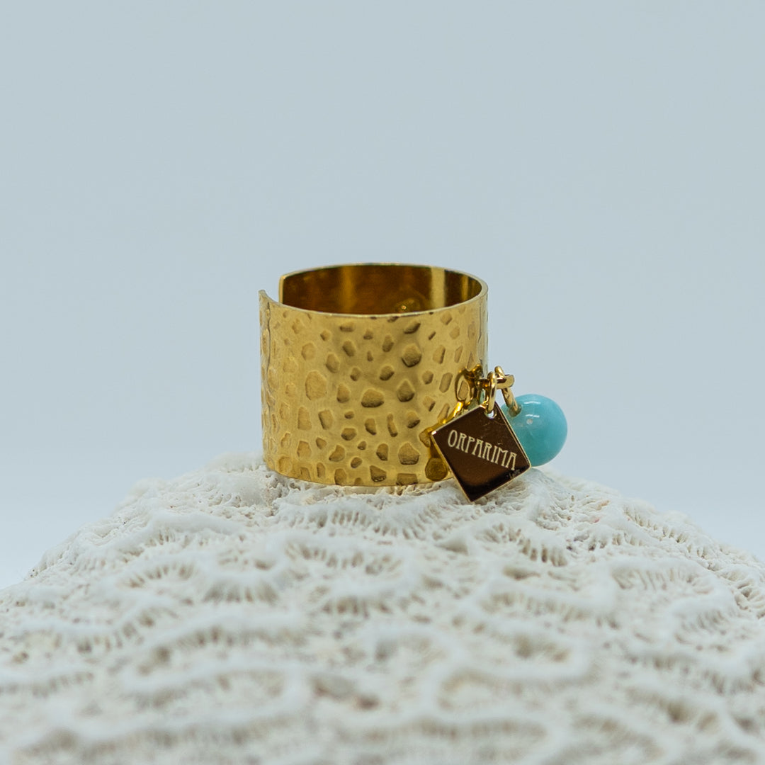 Bague prise en photo de coté posée sur un coquillage. Bague texturé en acier inoxydable 316L de couleur dorée, non fermée. Une breloque. avec l&