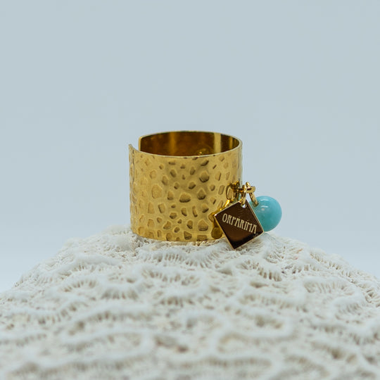 Bague prise en photo de coté posée sur un coquillage. Bague texturé en acier inoxydable 316L de couleur dorée, non fermée. Une breloque. avec l&