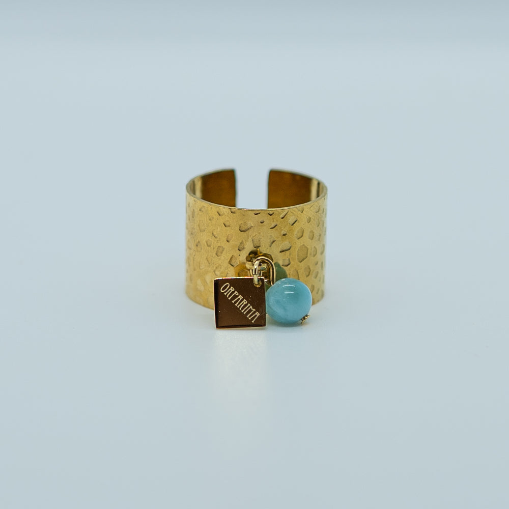 Bague large réglable de couleur dorée, faite en acier inoxydable 316L avec un anneau sur le dessus auquel sont accrochés une breloque orparima et une perle de larimar bleu de 6mm de diamètre.