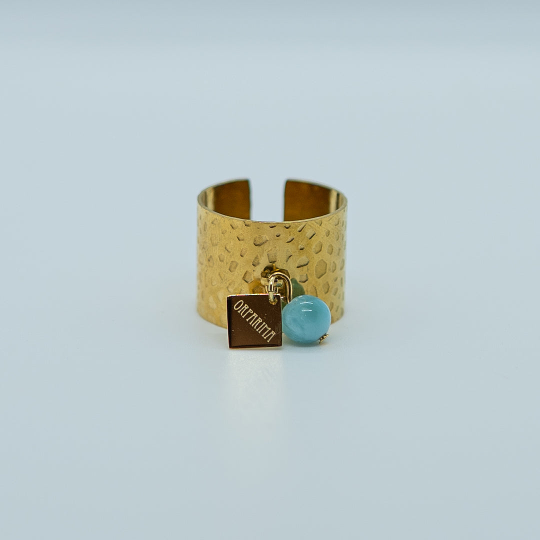 Bague large réglable de couleur dorée, faite en acier inoxydable 316L avec un anneau sur le dessus auquel sont accrochés une breloque orparima et une perle de larimar bleu de 6mm de diamètre.