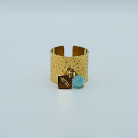 Bague large réglable de couleur dorée, faite en acier inoxydable 316L avec un anneau sur le dessus auquel sont accrochés une breloque orparima et une perle de larimar bleu de 6mm de diamètre.