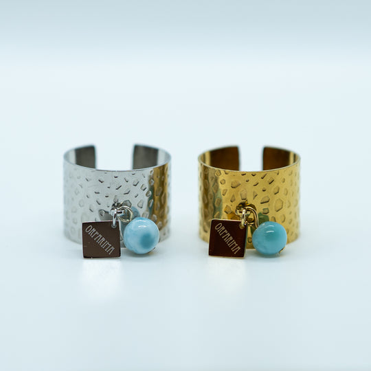 Deux bagues identiques vue de face sur fond blanc, les deux bagues sont texturés et possède un anneau sur le haut ou sont accrochées une breloque orparima et une perle de larimar de 6mm. La bague de gauche est en acier inoxydable 316L argent et la bague de droite est en acier inoxydable 316L doré. Les deux bagues sont réglables.