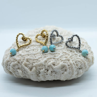 Deux paires de boucles d'oreilles en acier inoxydable 316L avec leur perle de Larimar pendante. Une paire de couleur argentée et l'autre de couleur dorée, toutes posée sur un coquillage.