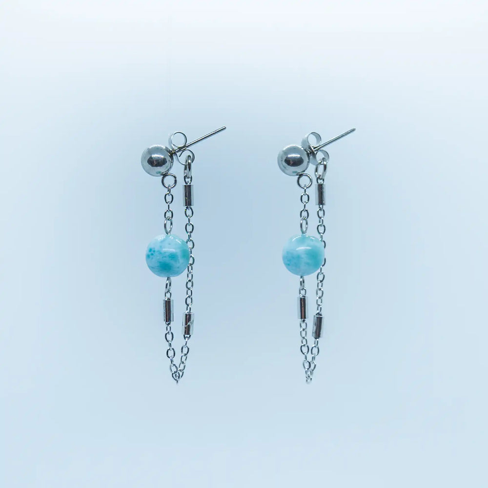 Paire de boucles d'oreille composées de la manière suivante : une chaîne relie la boucle d'oreille à une perle de Larimar, puis au fermoir. Ces boucles d'oreilles sont de couleur argentée et le Larimar est une pierre naturelle ne se trouvant qu'en République Dominicaine.