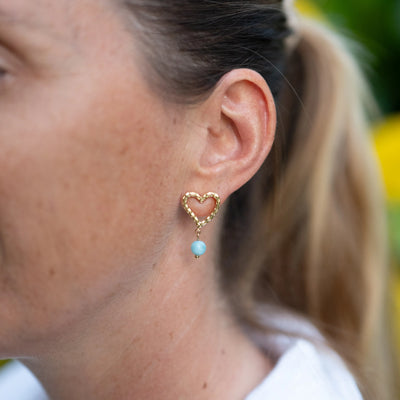Femme portant une boucle d'oreille en forme de cœur texturé comprenant une perle de Larimar en pendant. Boucle portée par une femme, bijou artisanal avec du Larimar.