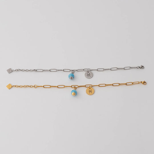 Beux bracelet ouvert en long et posés sur un fond blanc. Bracelet en acier inxydable 316L. Le bracelet du haut est couleur argent et celui du bas de couleur or. Une breloque orparima au bout de chaque chaine, un pendant signe astrologique et une perle de larimar.