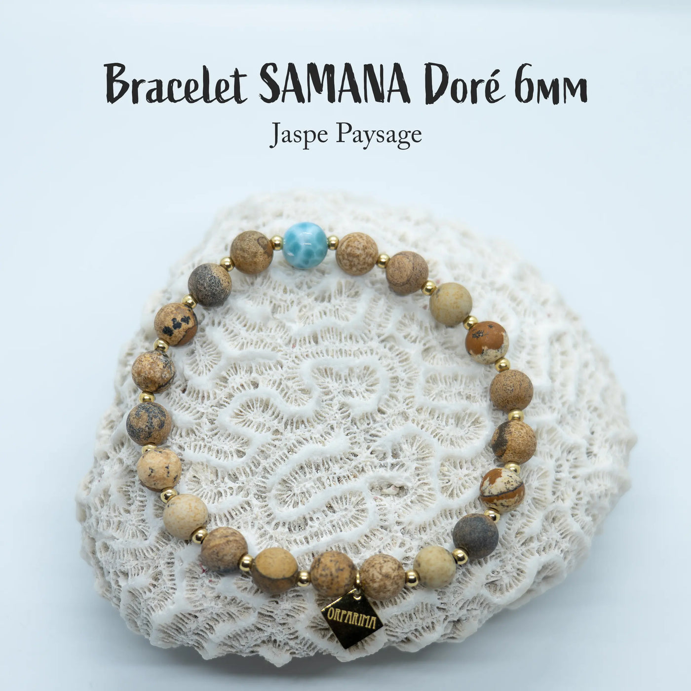 Bijou fait  à la main, posé sur un coquillage blanc, sur fond blanc. Ce bijou est un bracelet en fil élastique composé de perles de jaspe paysage et d'une perle de Larimar de République Dominicaine.