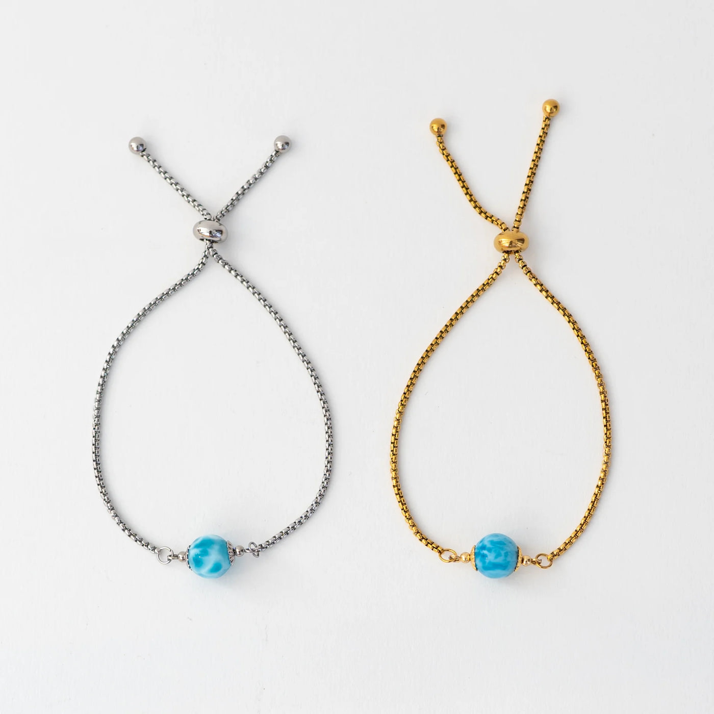 Photo de deux bracelets en acier inoxydable 316L, l'un est de couleur dorée l'autre est argenté. Ces deux bracelets sont montés avec une perle de larimar bleue de République Dominicaine, ils sont réglables grace à leur systède fermeture coulissant.