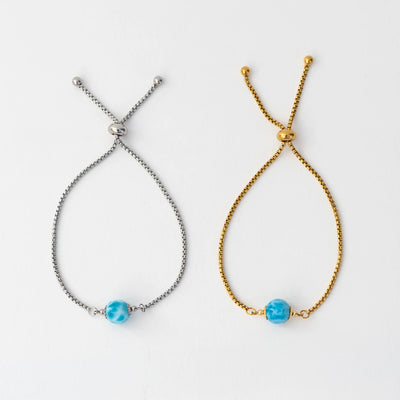 Photo de deux bracelets en acier inoxydable 316L, l'un est de couleur dorée l'autre est argenté. Ces deux bracelets sont montés avec une perle de larimar bleue de République Dominicaine, ils sont réglables grace à leur systède fermeture coulissant.