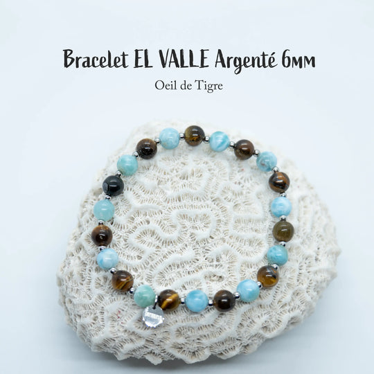 Bracelet avec une alternance de perles de Larimar et d’œil du tigre séparées par des petites perles en acier inoxydable argenté. Bijou posé sur un coquillage de type corail, fait avec un fil élastique.