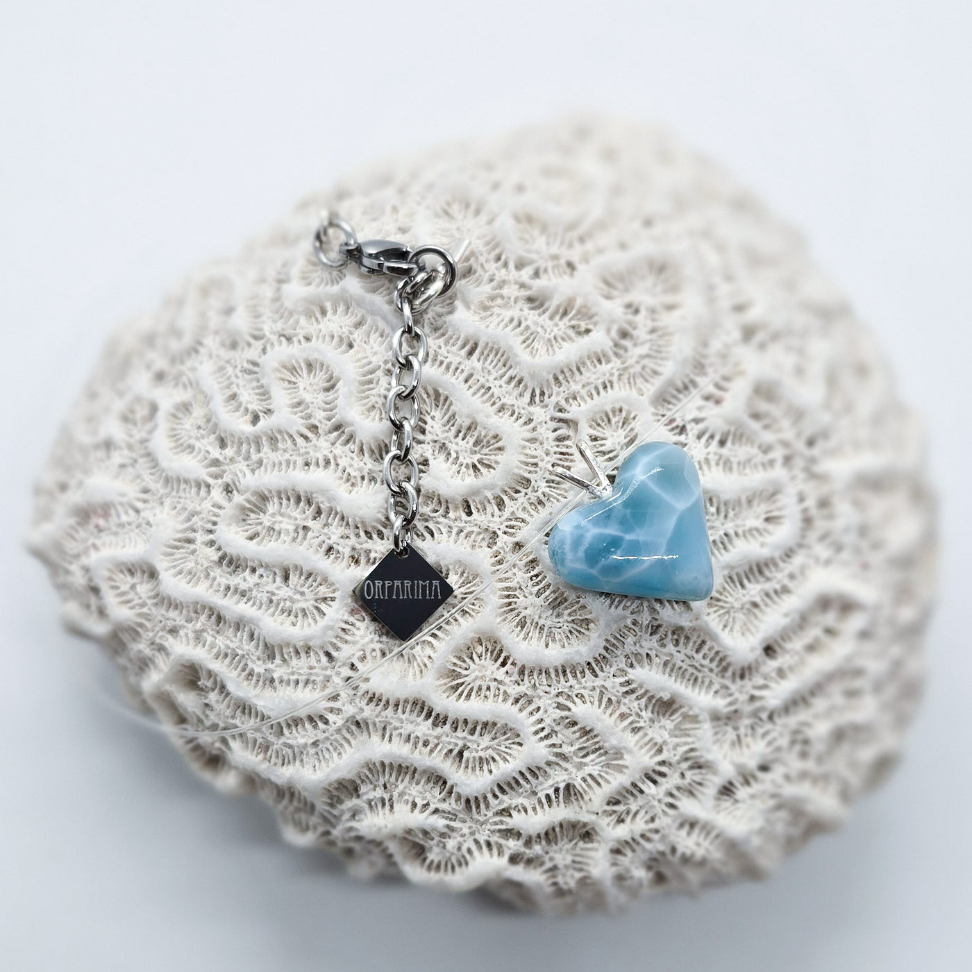 Pendentif en forme de cœur taillé dans la pierre naturelle de Larimar. Ce bijou est un collier en fil transparent avec des attaches en acier inoxydable chirurgical. Bijou posé sur un coquillage.
