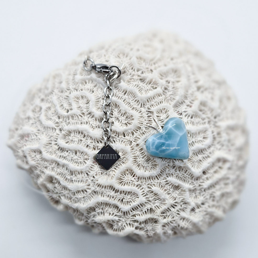 Petit cœur en Larimar posé sur un coquillage. Ce cœur est monté sur un fil transparent et possède une attache et une breloque Orparima en acier inoxydable 316L.