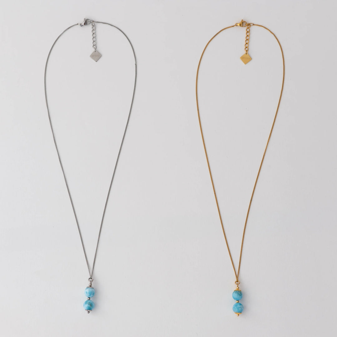 Deux colliers en acier inoxydable 316L argenté et doré posés à plat sur un fond blanc. Deux perles de larimar assemblées sur une tige sont les pendants de ces deux colliers. Extension de chaine avec une breloque orparima.