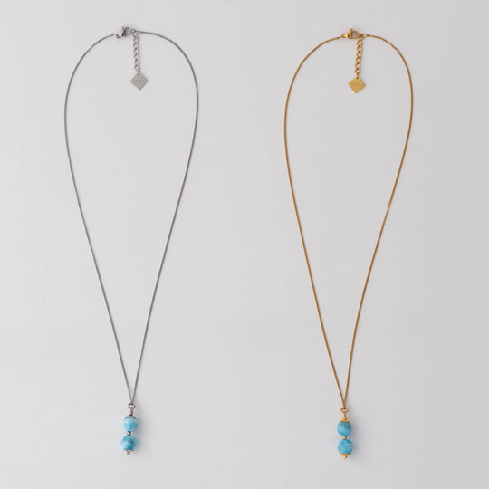 Deux colliers en acier inoxydable 316L argenté et doré posés à plat sur un fond blanc. Deux perles de larimar assemblées sur une tige sont les pendants de ces deux colliers. Extension de chaine avec une breloque orparima.