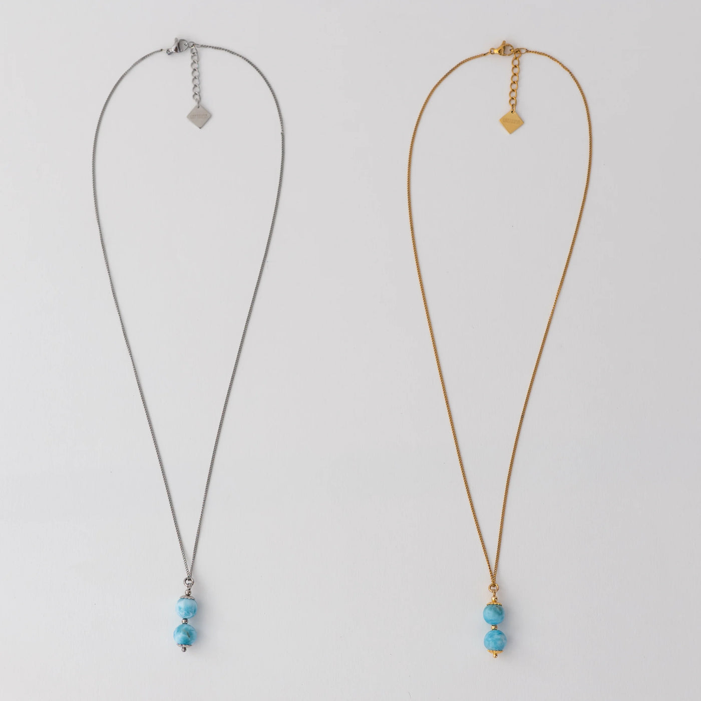Deux colliers en acier inoxydable 316L argenté et doré posés à plat sur un fond blanc. Deux perles de larimar assemblées sur une tige sont les pendants de ces deux colliers. Extension de chaine avec une breloque orparima.