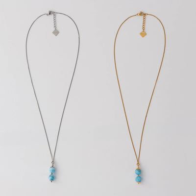 Deux colliers en acier inoxydable 316L argenté et doré posés à plat sur un fond blanc. Deux perles de larimar assemblées sur une tige sont les pendants de ces deux colliers. Extension de chaine avec une breloque orparima.