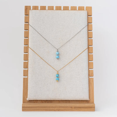 Deux colliers sur un présentoir. Le collier du haut est en acier inoxydable 316L argenté avec deux perles de larimar en pendant. L'autre collier est en acier inoxydable 316L doré avec aussi deux perles de larimar pendantes.