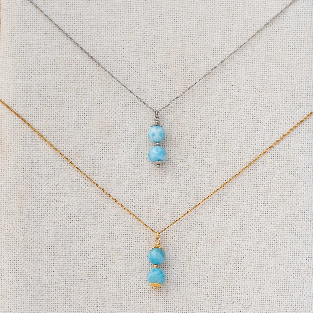 Photo de deux collier vue de prés, sur un présentoir. Chaque collier à deux perles de larimar comme pendant. Caqhue collier est en acier inoxydable, l'un est argenté et l'autre est doré.