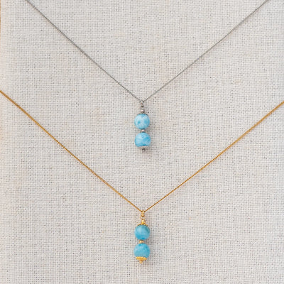 Photo de deux collier vue de prés, sur un présentoir. Chaque collier à deux perles de larimar comme pendant. Caqhue collier est en acier inoxydable, l'un est argenté et l'autre est doré.