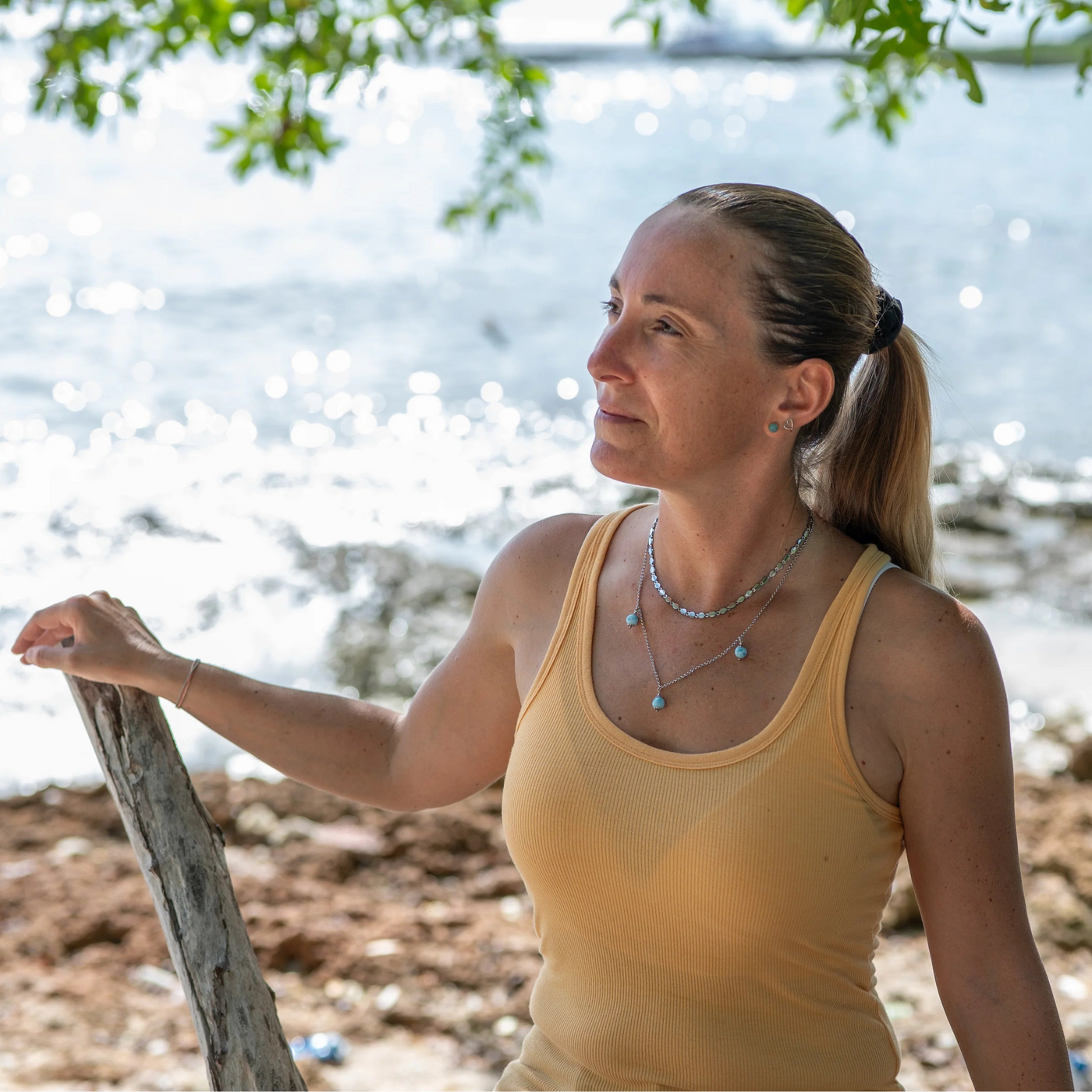 Femme portant un débardeur jaune posant devant une plage avec des rochers, seul la femme est nette sur la photo, l'arrière plan est flou. Elle porte un collier avec deux chaînes, chacune de couleur argentée, avec trois perles de larimar bleues. Les deux chaînes sont reliées mais de forme différentes.