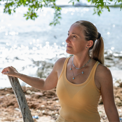 Femme portant un débardeur jaune posant devant une plage avec des rochers, seul la femme est nette sur la photo, l'arrière plan est flou. Elle porte un collier avec deux chaînes, chacune de couleur argentée, avec trois perles de larimar bleues. Les deux chaînes sont reliées mais de forme différentes.
