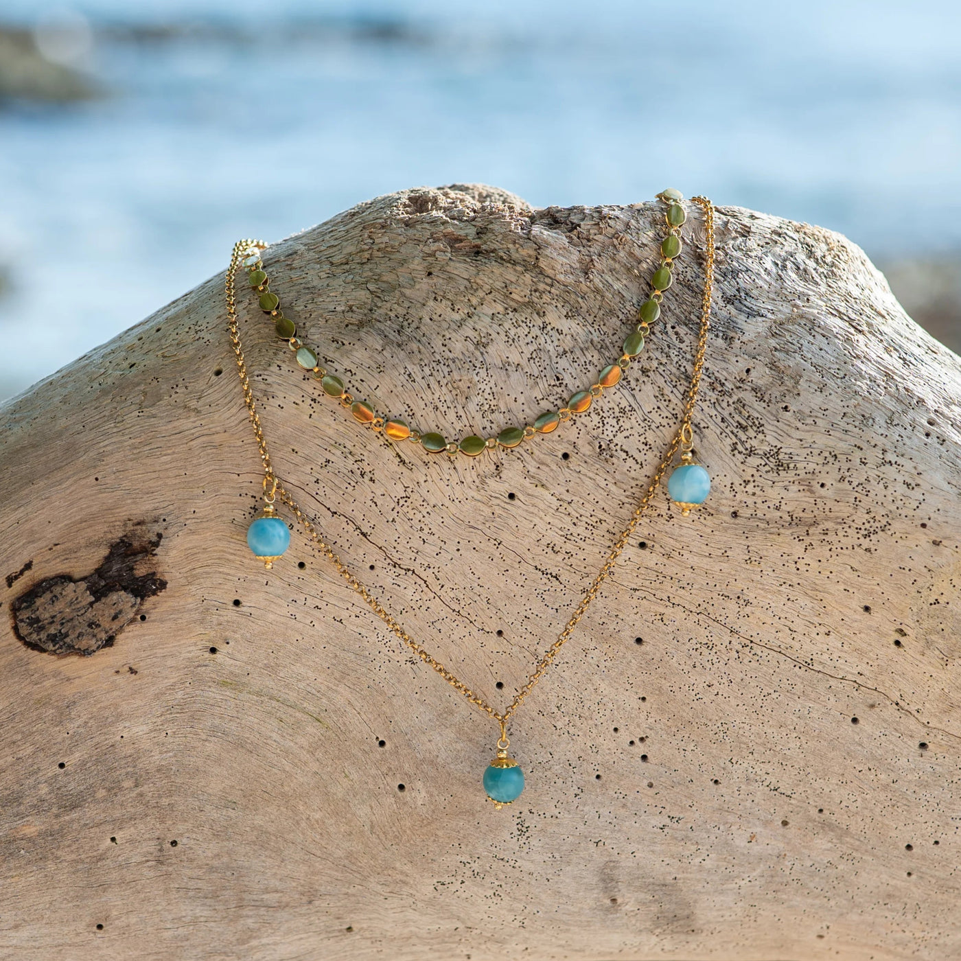 Collier avec deux chaines de forme et de longueur différentes, avec trois perles de larimar de différents bleu, posé sur un morceau de bois flotté avec la mer en arrière plan.