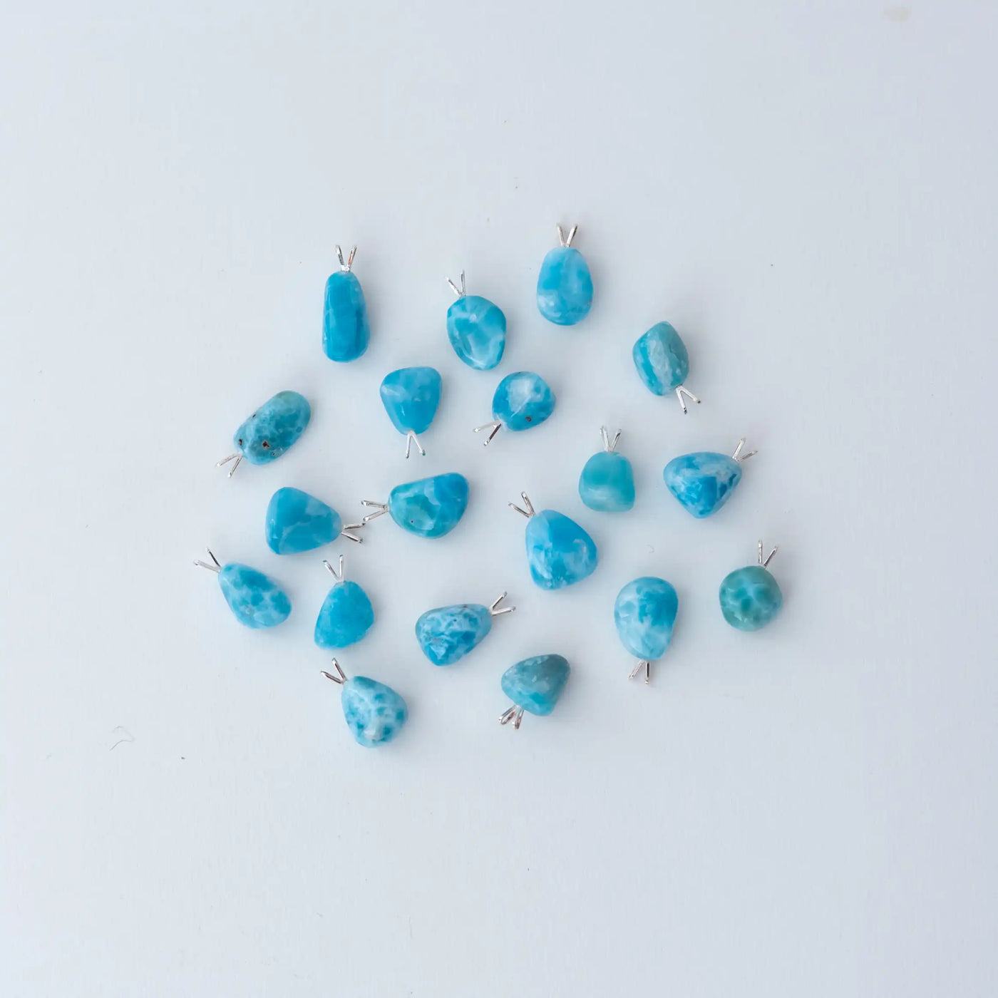 dix-neuf cabochons différents en terme de taille et de couleur, tous fait en Larimar, son posés sur un fond blanc et vue de dessus. Ces cabochons font environ deux centimètres de longueur et peuvent être montés en collier.