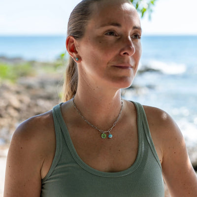 Photo du haut du corps d'une femme prise jusqu'au sommet du crane, posant devant une mer en arrière plan et portant un collier en acier inoxydable 316L de couleur argent, avec une perle de larimar et un signe astrologique en pendants.