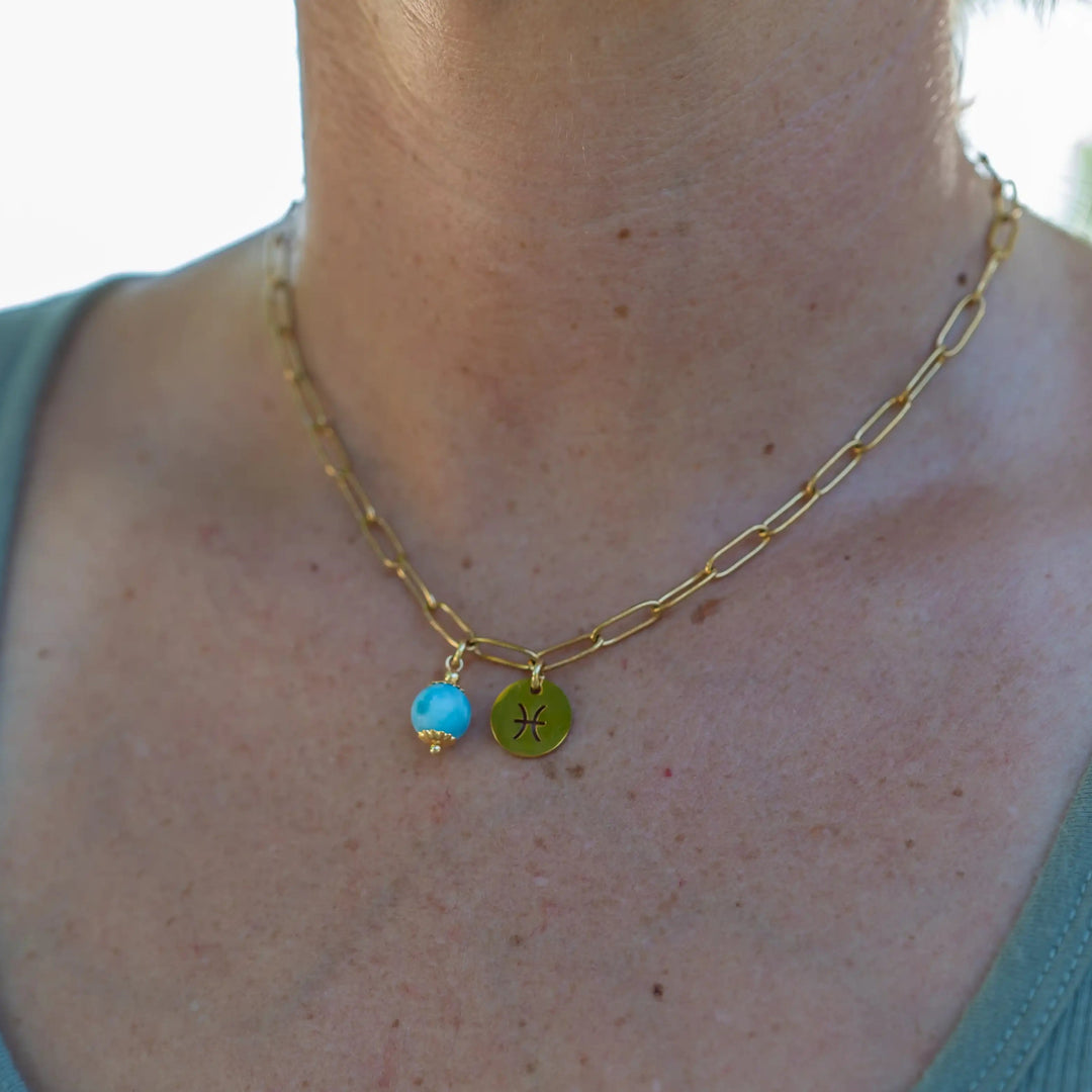 Collier de couleur doré porté par une femme dont on ne voit que le torse et le cou. Le collier est en acier inoxydable et comprend deux pendants, un signe astrologique et une perle de larimar de République Dominicaine. 