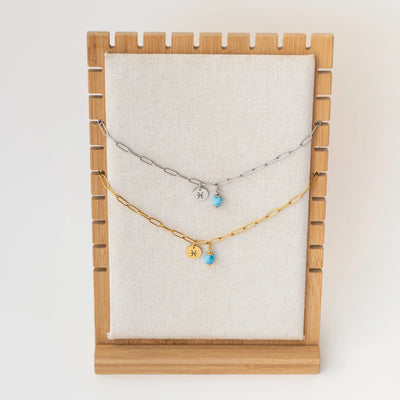 Photo de deux colliers avec des mailles larges, suspendu sur un présentoir, l'un des bracelet est en acier inoxydable 316L argenté et l'autre est en acier inoxydable doré. Une perle de larimar et un signe astrologique son attachés à la chaineen son milieu.